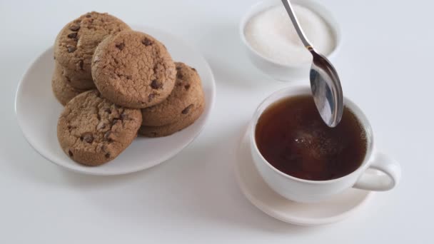 Tillsätt socker till kopp te eller kaffe med cookies på vitt köksbord. Närbild. — Stockvideo