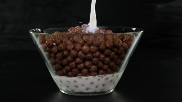 Verter la leche y las bolas de cereales de chocolate en un recipiente de vidrio sobre fondo negro. De cerca. . — Vídeo de stock