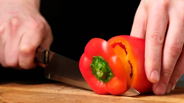 Knife cuts the red pepper in kitchen on wooden board. Close up.