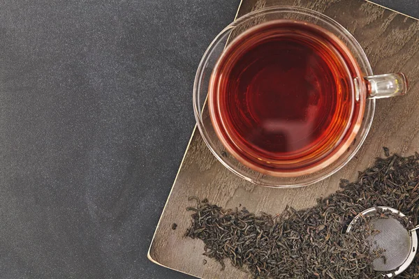 Glass cup of hot tea on dry black tea leaves on wooden backgtound. Top view. — Stock Photo, Image