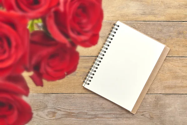 简朴的棕色木桌上的空白笔记本和红玫瑰的顶部视图. — 图库照片