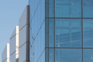 Büyük bir şehrin merkezindeki modern bir yüksek katlı ofis veya ticari binanın ön cephesinde cam şeffaf pencereler mavi gökyüzünü yansıtır.