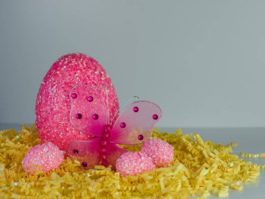 Büyük pembe Paskalya yumurtası 3 küçük pembe Paskalya yumurtasıyla çevrili sarı kağıt parçalarından oluşan bir yatakta oturmuş değerli taşlı pembe bir ağ kelebeğiyle..