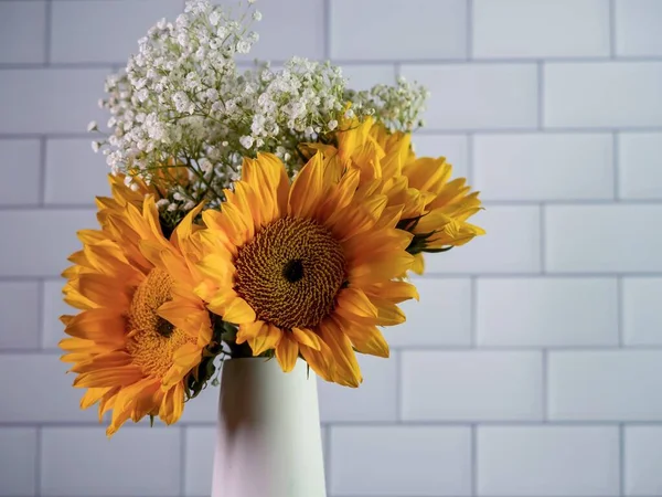 Yellow sunflowers with white babies breath floral arrangement in a ceramic white vase with a white subway tile background.  Colorful, fresh cut flowers, home decor.