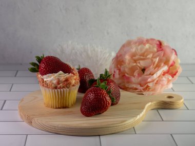 Krem şantili sarı kek taze çilekle süslenmiş kek ve daha fazla taze çilek ve arkasında pembe ve beyaz suni bir çiçek olan ahşap kesme tahtasının üzerinde kek parçaları..