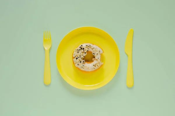 stock image Bite donut on yellow plate with knife and fork on green background. Minimal creative unhealthy food concept.