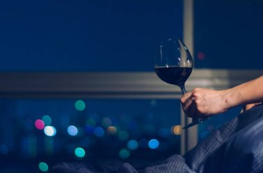 Yatağın üzerinde kırmızı şarap kadehi tutan bir adam. Şehir ışıklarından renkli bir bokeh taşıyor..