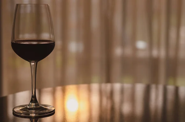 A glass of red wine on table in the room with sunset light on curtian background.