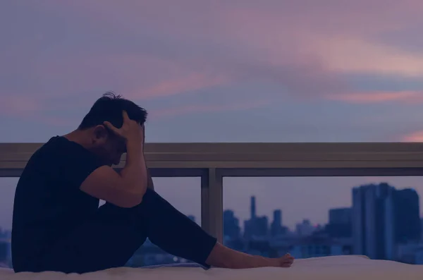 A person sitting and feeling depressed on bed with city view in dusk moment. Stay home, depression and loneliness concept.