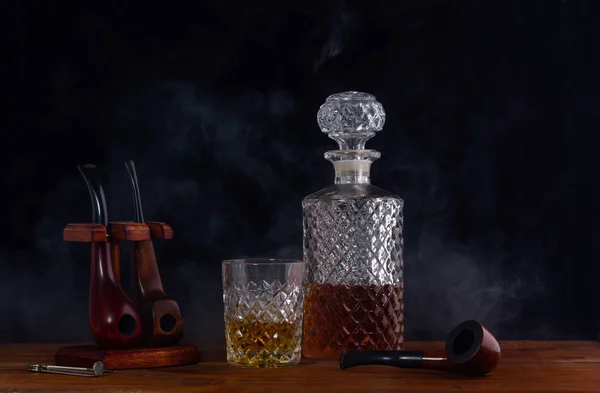 Carafe and a glass of whiskey beside the smoking tubes standing on the table shrouded in smoke from the tube — Stock Photo, Image