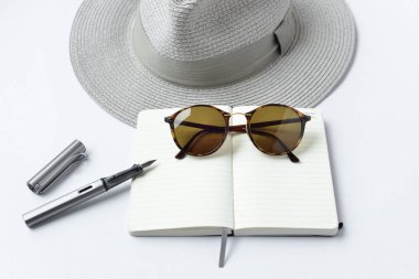 Güneş gözlüğü, defter, kalem ve beyaz zemin üzerine şapka