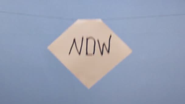 Мужчина вешает белый лист бумаги с черной надписью "сейчас" на синем фоне — стоковое видео