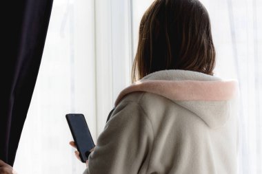 Evdeki pencerede elinde telefon olan kız. Koronavirüs salgınından dolayı karantina sırasında bir kız pencereden dışarı bakar. İzolasyon ve yalnızlık.