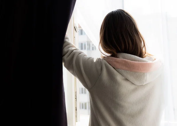 The girl at home opens the window for airing.A girl during quarantine due to the coronavirus pandemic looks out through a window. Isolation and self-isolation.