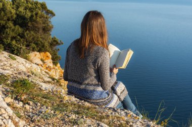 Uçurumun kenarında kitabı olan bir kız. Doğada uzak bir çalışma. Doğada eğitim. Doğayı deniz manzaralı okumak. Doğada yalnızlık. Kişisel izolasyon ihlali.