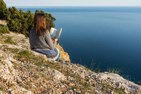 A girl with a book on the edge of a cliff. Remote work in nature. Training in nature. Reading in nature with a sea view. Self-isolation in nature. Violation of self-isolation.
