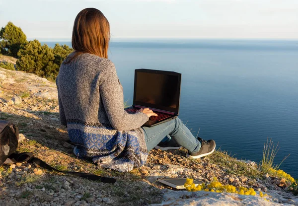 A girl is working on a laptop at the top of a mountain. Remote work and training in nature. Sea view. Self-isolation in nature. Violation of self-isolation. Isolated from other people.