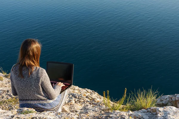 A girl is working on a laptop at the top of a mountain. Remote work and training in nature. Sea view. Self-isolation in nature. Violation of self-isolation. Isolated from other people.