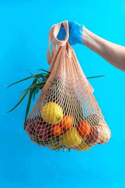 String bag with products on a blue background. A girl holds a bag of groceries. Food delivery. A volunteer delivers food to the coronavirus. Fresh food in an eco bag. Eco-friendly lifestyle.