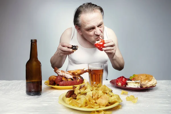 Older man in a-shirt eating lot of grilled meat, sweet sugar cakes, potato chips and beer.