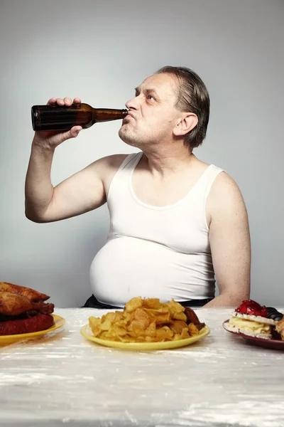 Older man in a-shirt drinking beer and eating lot of junk food.