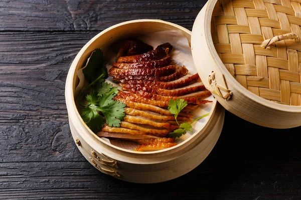 Sliced Peking Duck in bamboo steamer with fresh cilantro on black burned wooden background