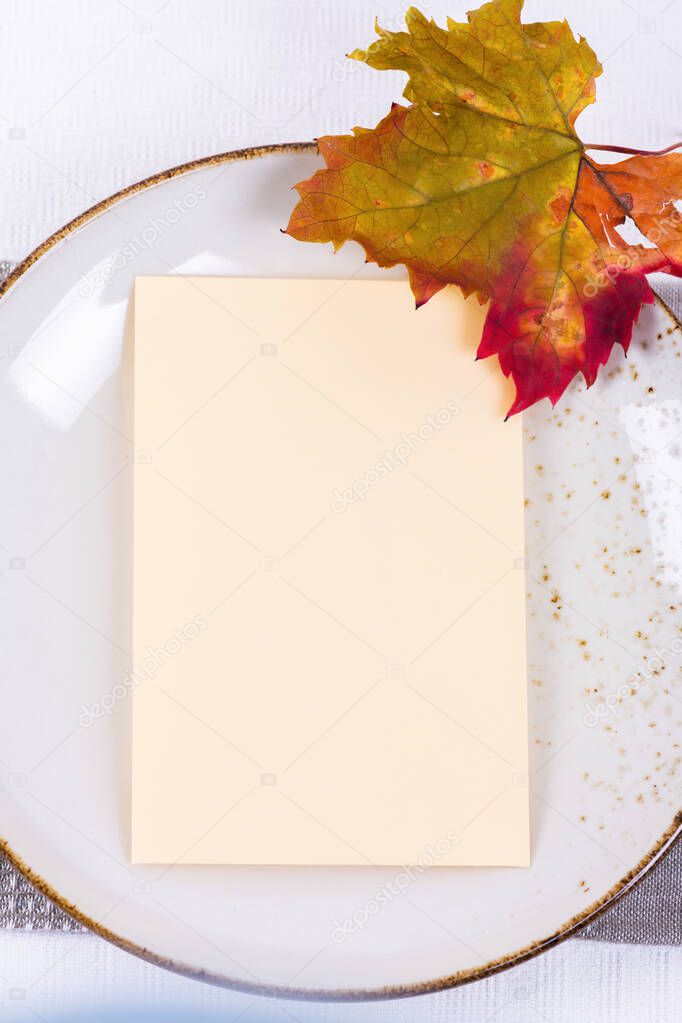 Blank card mock up invitation or menu on porcelain plate with festive fall table setting with colorful autumn leaves. Minimal restaurant menu concept. Flat lay, top view