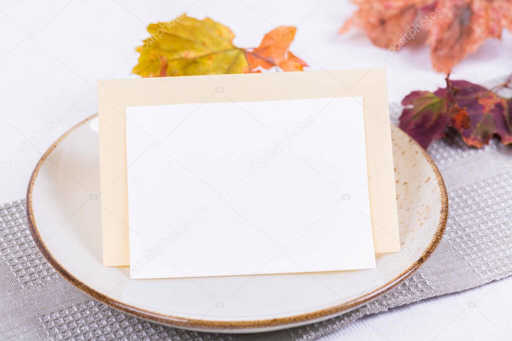 Blank card mock up invitation or menu on porcelain plate with festive fall table setting with colorful autumn leaves. Minimal restaurant menu concept. Flat lay, top view
