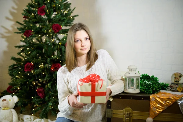 Menina senta-se perto de um abeto de Natal e mantém em suas mãos um presente — Fotografia de Stock