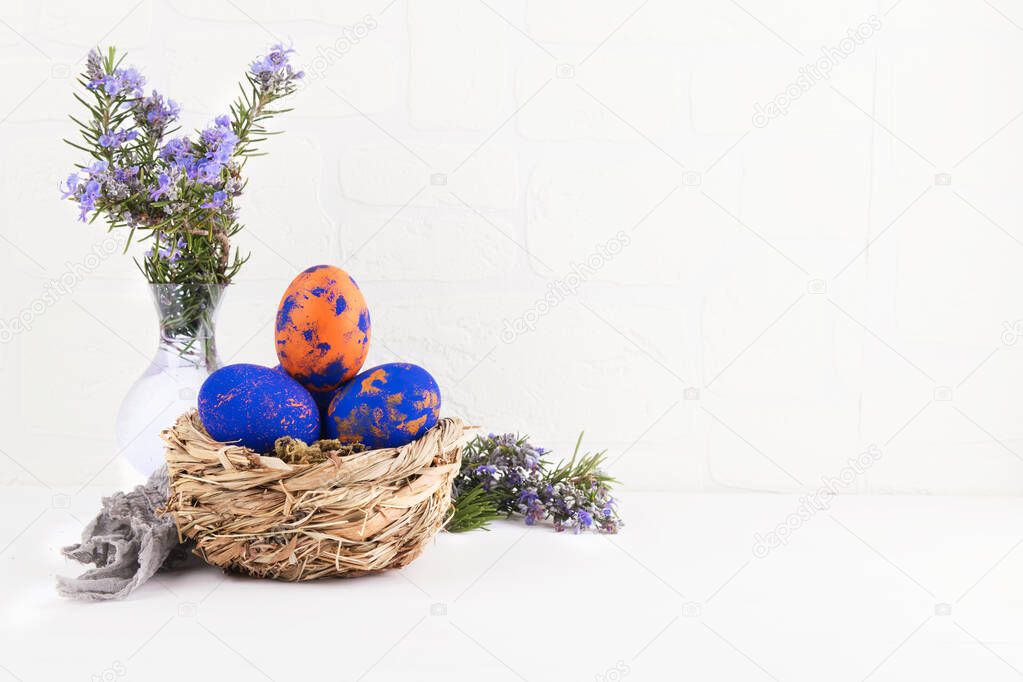 A nest with with classic blue and orange speckled easter eggs, a brunch of rosemary flowers i layng on the table on a withe wall background. Concept of Easter holyday and spring break