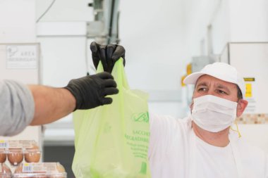 Avetrana, İtalya, Mart 19, 2020. Satıcı, Coronavirus salgını sırasında tıbbi maske ve eldiven takarak bir müşteriye et servisi yapıyor. Alışveriş, Covid-19 pandemisi, karantina