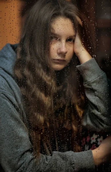 Young girl stay at home isolation, during quarantine for stave off preading of disease coronavirus, near a window on which raindrops. Quarantine mood depression, fear, loneliness, pandemia Covid 19