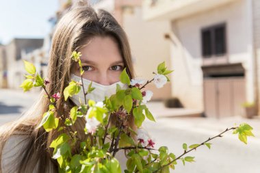 Tıbbi maske takan kız umutla bakıyor ve elinde kayısı dalları tutuyor. Karantinanın sona ermesi ve koronavirüsü yenmek ve normal hayata dönmek.