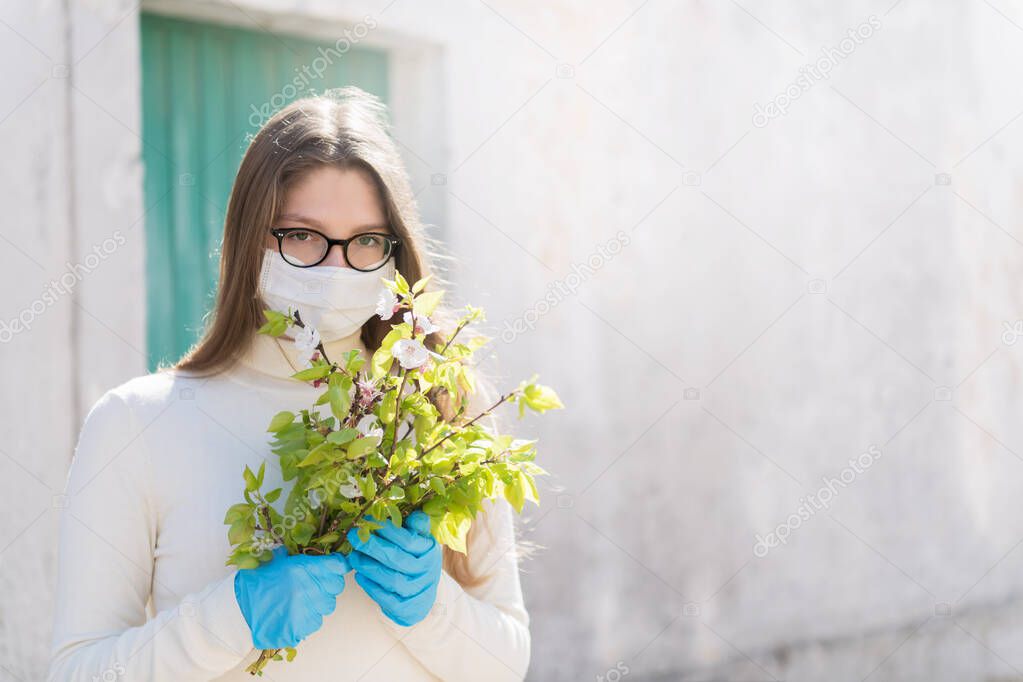 Girl wearing medical mask, is looking with hope and holding a branch of blooming twigs of apricot. Concept of end of quarantine and win over coronavirus and returning to normal life, copy space