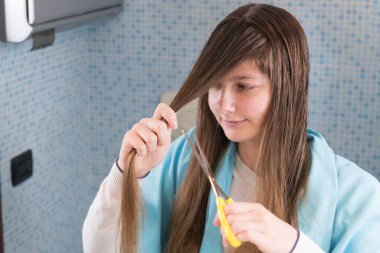 Bir kız, Coronavirus 'u kapatırken saçlarını kesmek için elinde makasla aynaya bakıp şaşkın ve komik bir yüz ifadesi takınıyor. Coperid-19 sırasında saç kesimi, evde kal, seçici odaklanma