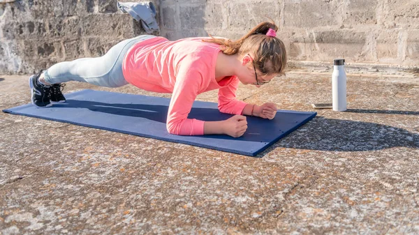 Genç kız Coronavirus karantinası sırasında evinin çatısındaki hasırın üzerinde kalas yapıyor. Sağlıklı kalmak için kadın yoga eğitimi. ev işleri, karantina sporu, ev antrenmanı
