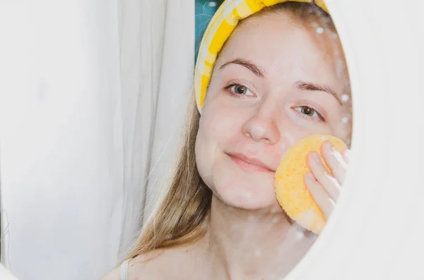 A European girl is applying Korean cosmetics to her face mask. Cleanses pores, grease, acne, wrinkles. Close-up face of a girl with skin problems. Mousse, spray, face scrub and face wash.Yellow Sponge — Stock Photo, Image