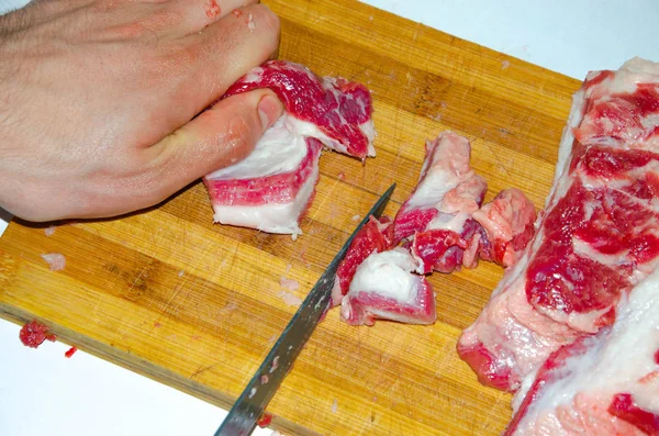 Rått kött på tavlan. En man skär fläskkött i bitar med kniv. Kött med bacon. Sjukdomar i rått kött. Feta händer från kött — Stockfoto