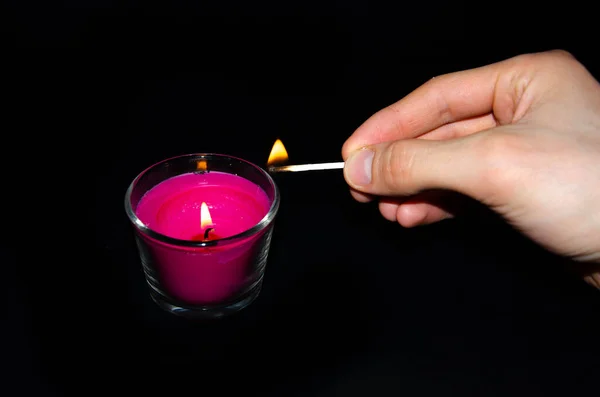Bright pink candle in a glass jar on a black background. The candle is burning. Redeemed. A hand sets fire to a candle with matches. Put out the candle with your fingers, hands.
