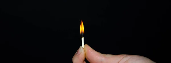 A hand holds a burning match on a black background. A wooden match burns in the hands of a macro. Igniting a match on a box. Smoke from a match. Flash