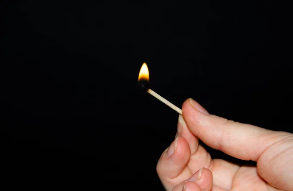 A hand holds a burning match on a black background. A wooden match burns in the hands of a macro. Igniting a match on a box. Smoke from a match. Flash