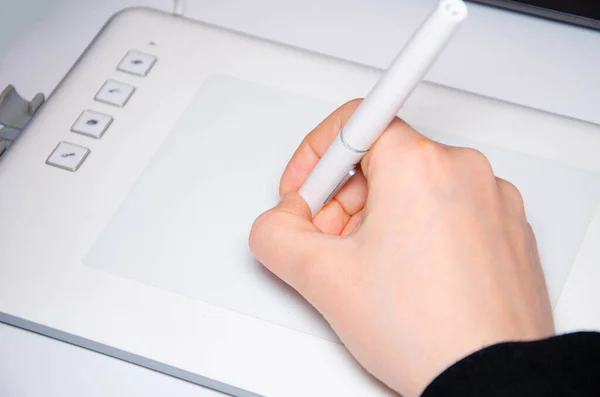Female hands work on a graphic tablet. Hand holds stylus pen and draws. White graphic tablet. The work of a graphic designer. Girl works on a tablet connected to a laptop. Rear view from behind