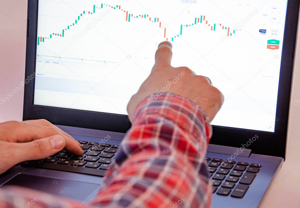 Trading on the exchange. Men's hands in a plaid shirt behind a laptop show on the graph. On-screen rates, promotions. Wins are losses. Part of the back and head is visible Focus on laptop and graphics
