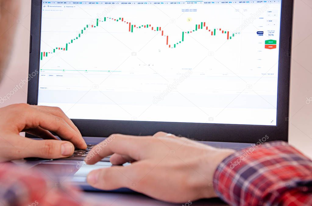 Trading on the exchange. Men's hands in a plaid shirt at a laptop. On-screen rates, promotions. Schedule. Wins are losses. Part of the back and head is visible. Focus on laptop and graphics