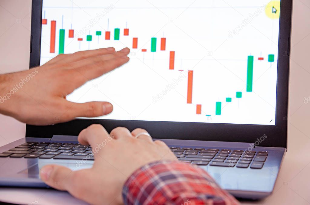 Trading on the exchange. Men's hands in a plaid shirt behind a laptop show on the graph. On-screen rates, promotions. Wins are losses. Part of the back and head is visible Focus on laptop and graphics
