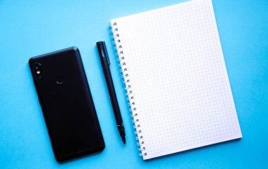 Mavi arka planda kare şeklinde bir defter. Siyah kalem, defter ve akıllı telefon. Not defteri, planlayıcı. Mesaj için yer. Sarmalı bir defter.