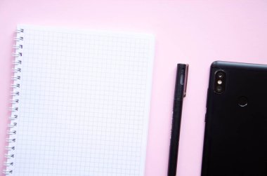 Pembe arka planda kare şeklinde bir defter. Siyah kalem, defter ve akıllı telefon. Not defteri, planlayıcı. Mesaj için yer. Sarmalı bir defter.
