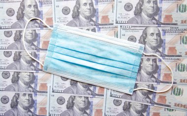 100 dolarlık banknotların arka planında mavi tıbbi maske. Uzayı kopyala COVID-19