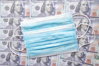 100 dolarlık banknotların arka planında mavi tıbbi maske. Uzayı kopyala COVID-19