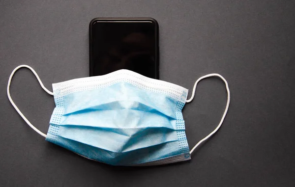 Siyah bir akıllı telefondaki mavi tıbbi maske. Koruma, akıllı telefonun dezenfeksiyonu. Maske, mavi, siyah, pembe, sarı arka plandaki akıllı telefondan giyinmiş. Uzayı kopyala COVID-19